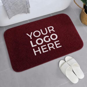 May include: A rectangular deep red bath mat with rounded corners featuring the white text "YOUR LOGO HERE" printed in the center. The mat has a textured, plush surface and is positioned on a light gray tiled floor next to white slide sandals.