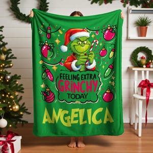 May include: Green Christmas blanket featuring the Grinch holding a candy cane, wearing a Santa hat and red coat. The blanket has the text "Feeling Extra Grinchy Today" and the name "Angelica". Decorative Christmas ornaments are also featured.