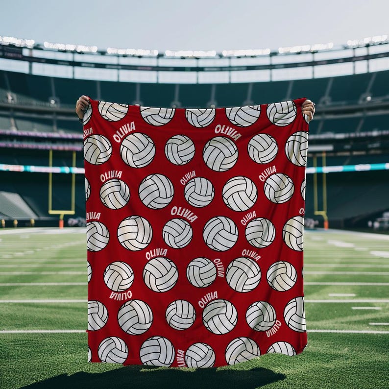 May include: A red blanket featuring a pattern of white volleyballs and the name "Olivia" in white. The blanket is held up against a backdrop of a football field and stadium.