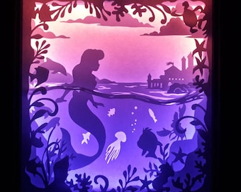 May include: A purple and pink paper cut-out art piece depicting a mermaid swimming in the ocean. The mermaid is silhouetted against a pink and purple background. The ocean is filled with sea life, including fish, jellyfish, and seaweed. The scene is framed by a black frame.