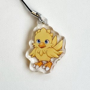 May include: A clear acrylic charm featuring a yellow cartoon chick with blue eyes and orange feet. The charm is attached to a black cord with a silver-colored clasp. The chick has a detailed design and is set against a white background.