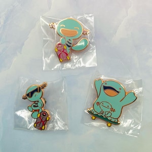 May include: Three enamel pins featuring cartoon axolotls on skateboards. The pins are in clear plastic packaging. The axolotls are light blue with gold outlines and orange mouths. The skateboards are pink, red, and green.