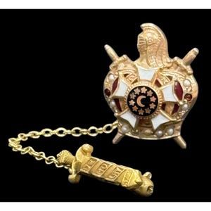 May include: A gold-tone metal lapel pin featuring a knight's helmet above a cross with white and red enamel accents, a central black circle with a crescent moon and stars, and a chain connecting to a bar pin engraved with the text "MO LY".