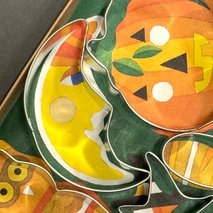 K&ouml;nnte beinhalten: Ein Set von Halloween-Ausstechern. Die Formen umfassen einen l&auml;chelnden Mond, einen K&uuml;rbis und eine Eule. Die Ausstecher sind aus silberfarbenem Metall gefertigt und mit farbenfrohen Papierdesigns gef&uuml;llt. Der Hintergrund ist gr&uuml;n.