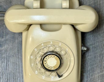 Könnte beinhalten: Ein beigefarbenes Wählscheibentelefon mit einem weißen Zifferblatt und schwarzen Zahlen. Das Telefon hat ein beigefarbenes Hörer und ist an einer Wand montiert.