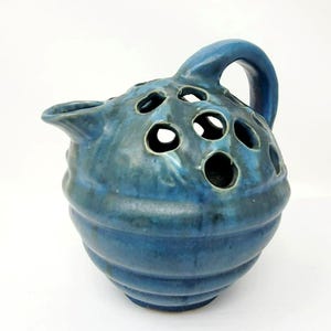 May include: A blue ceramic vase with a handle and a spout. The vase has a series of circular holes on the top and a ribbed design on the body. The glaze has a mottled appearance.