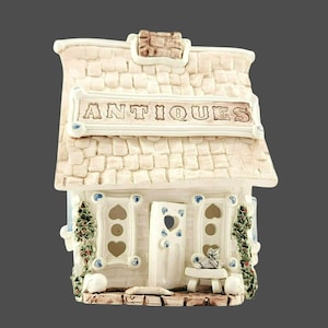 May include: A miniature ceramic antique shop figurine. The shop has a white exterior with a textured roof and a sign that reads "ANTIQUES". The windows and door have heart-shaped cutouts. A small cat sits on a bench outside the door.