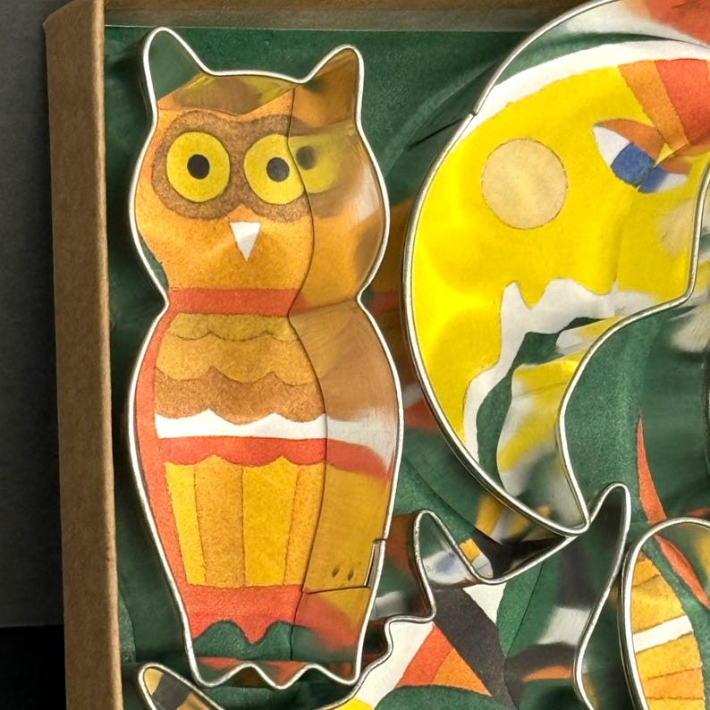 K&ouml;nnte beinhalten: Ein Set bunter, eulenformiger Ausstecher mit einem Aquarelldesign. Die Eule hat gelbe Augen, einen wei&szlig;en Schnabel und orangefarbene und gelbe Federn. Die Ausstecher befinden sich in einer Pappschachtel mit gr&uuml;nem Hintergrund.