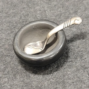 May include: A small, round, black ceramic bowl with a silver spoon resting inside. The spoon has a decorative handle and a curved bowl. The bowl and spoon are set against a gray background.