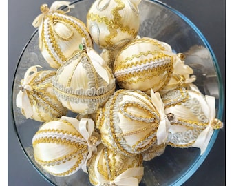 Può includere: Una collezione di ornamenti natalizi color crema con finiture decorative dorate e argentate, esposti in una ciotola di vetro trasparente. Ogni ornamento è adornato con un fiocco di raso e dettagli intricati, creando un'elegante esposizione festiva.