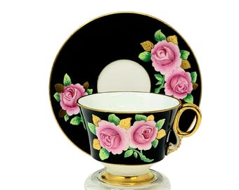 May include: A black teacup and saucer set with a gold rim and pink rose floral design. The cup and saucer are decorated with pink roses and green leaves on a black background.