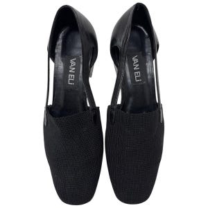 May include: A pair of black leather heeled shoes with a mesh fabric toe. The shoes have a cut-out design on the sides and the brand name "VAN ELI" is printed on the inside of each shoe.