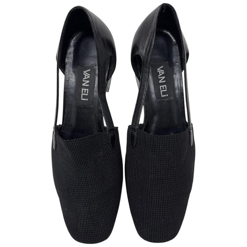 May include: A pair of black leather heeled shoes with a mesh fabric toe. The shoes have a cut-out design on the sides and the brand name "VAN ELI" is printed on the inside of each shoe.