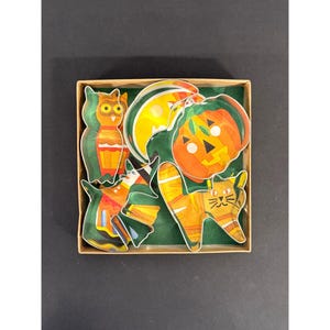 K&ouml;nnte beinhalten: Ein Set von Halloween-Papierausschnitten in einer Box. Die Ausschnitte umfassen eine Eule, eine Hexe, eine Katze und zwei K&uuml;rbisse. Die Eule ist orange und gelb, die Hexe ist orange und blau, die Katze ist orange und gelb und die K&uuml;rbisse sind orange und gelb.