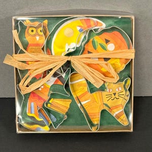 K&ouml;nnte beinhalten: Ein Set von Halloween-Untersetzern in einer transparenten Box, gebunden mit Bast. Die Untersetzer zeigen eine Eule, einen Halbmond, einen K&uuml;rbis und eine Katze. Die Untersetzer sind orange, gelb und gr&uuml;n mit schwarzen Umrissen.