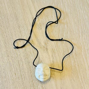 May include: A white ceramic pendant shaped like a face with a smiling expression, hanging from a black cord.