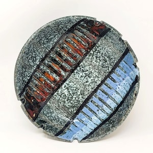 May include: A round ceramic ashtray with a speckled grey glaze. The ashtray has three bands of colour: a blue band, a red band, and a black band. The bands are decorated with lines and geometric patterns.