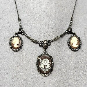 May include: A silver-toned necklace featuring three pendants. Two oval pendants display cameo profiles, while the central pendant showcases a white flower design on a brown background. The necklace has a delicate chain and decorative accents.