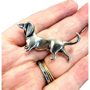 May include: A silver-coloured dachshund dog brooch. The brooch depicts a dog with its head raised and tail curled. The brooch is made of a shiny metal and has a pin clasp on the back.