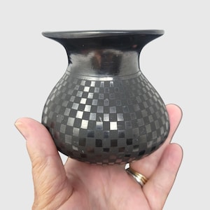 May include: A small, rounded black ceramic vase featuring a polished finish and a repeating checkerboard pattern across its body. The vessel has a wide, flared rim and a smooth, dark surface that reflects light, creating a contrast between the matte and glossy squares.