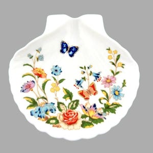 May include: White seashell-shaped dish with a colourful floral and butterfly design. The dish features a variety of flowers in shades of pink, blue, yellow, and orange, along with two butterflies. The edges of the dish are scalloped.