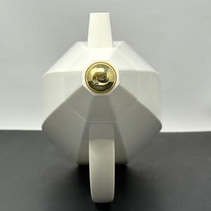 Peut inclure: Une théière blanche géométrique dotée d'un corps anguleux à facettes et d'un bouton métallique doré circulaire proéminent centré sur le couvercle. La théière possède une anse incurvée partant du bas et un bec verseur vertical au sommet.