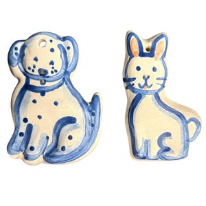 May include: Two ceramic ornaments shaped like animals, featuring a cream-colored base with hand-painted blue outlines and details. One ornament depicts a spotted dog, while the other represents a cat with pointed ears, each with a hole for hanging.