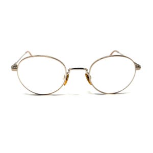 May include: A pair of round eyeglasses with a thin, metallic silver-toned frame. The glasses feature a slim bridge, clear lenses, and visible amber-colored nose pads attached to the inner rims, providing a simple and functional eyewear design.