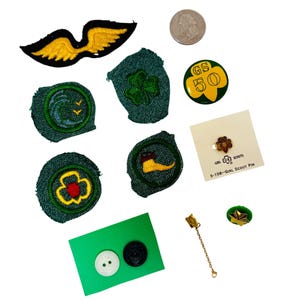 May include: A collection of Girl Scout badges and pins, including a green and yellow badge with the text "GS 50", a green badge with a four-leaf clover, a green badge with a wave and birds, a green badge with a flower, a green badge with a boot, a gold pin with a star, a gold pin with a chain, and a green and gold pin with the text "Girl Scouts 9-106-Girl Scout Pin".