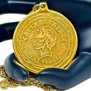 May include: A gold-colored necklace with a round pendant featuring the words "Caesar's Palace Las Vegas".