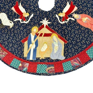 May include: A circular fabric tree skirt featuring a dark blue background with a gold star pattern. The center displays a nativity scene with a stable, figures, and angels. The border consists of alternating red and teal patterned fabric squares.