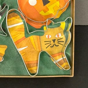 K&ouml;nnte beinhalten: Ein Set von Halloween-Ausstechern in einer Pappschachtel. Die Ausstecher haben die Form einer Katze, eines K&uuml;rbisses und eines Hexenhuts. Der Katzenausstecher ist orange und gelb mit schwarzen Details. Der Hintergrund ist gr&uuml;n.