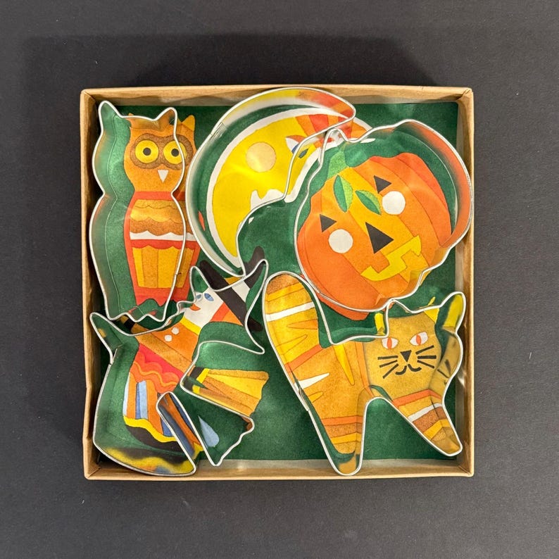 K&ouml;nnte beinhalten: Eine Sammlung von Halloween-Papierauschnitten in einer Pappschachtel. Die Ausschnitte umfassen eine Eule, einen K&uuml;rbis, eine Katze, eine Hexe und einen Halbmond. Die Farben sind Orange, Gelb, Gr&uuml;n und Schwarz.