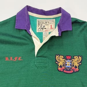 May include: Green rugby shirt with a purple collar, a white under-collar, and a crest with two golden dragons on the right side. The crest has a blue shield with a white "R" and the words "Ralph Lauren Rugby" below. The shirt also has the text "R.L.F.C." on the left side and a tag that says "Rugby Ralph Lauren Established 2004" on the upper right side.