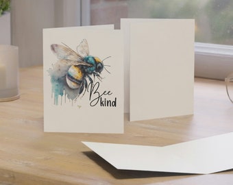 May include: A white card with a watercolor illustration of a bee with yellow and black stripes and blue wings. The text "Bee kind" is written in black below the bee.
