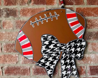 May include: A brown football-shaped wooden sign with white stitching and red and gray stripes. A large black and white houndstooth bow is attached to the bottom. The sign is hanging on a brick wall.