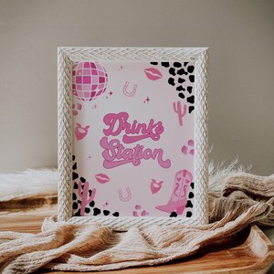 May include: White framed sign with a pink background and black and white cow print accents. The sign says "Drinks Station" in pink script with a pink disco ball, pink cowboy boots, and pink horseshoes.
