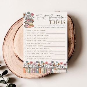 May include: A First Birthday Trivia game card with floral and book illustrations. The card features fill-in-the-blank questions about the birthday girl, such as her nickname, favorite toy, and first word. The card is on a wooden surface.