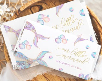 Op de afbeelding: Twee witte snoepreepverpakkingen met zeemeerminstaart- en bellenontwerpen. De verpakkingen hebben een gouden tekst die luidt "our little mermaid" en "MADDISON IS TWO!". De verpakkingen liggen op een houten kom.