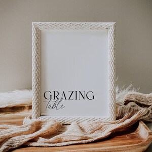 Puede incluir: Marco de madera blanco con un paspartú blanco y el texto "GRAZING table" en letras negras. El marco está sobre una superficie de madera marrón con una tela beige drapeada sobre él.
