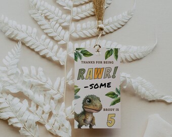 Puede incluir: Una etiqueta de favor de fiesta de dinosaurio rosa y verde con el texto "Gracias por ser RAWWR! -SOME" y una ilustración de dinosaurio verde. La etiqueta también incluye el texto "Brody tiene 5 años".