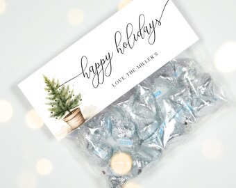May include: A white gift bag with a watercolor illustration of a small potted Christmas tree. The bag has a label that reads "happy holidays" in cursive script and "LOVE THE MILLERS" in a smaller font. The bag is filled with silver foil-wrapped chocolate kisses.