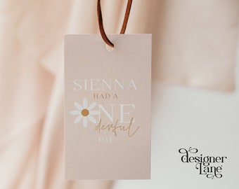 May include: A pink tag with the text "Sienna had a one derful day" in white and gold lettering. A white daisy with a gold center is next to the word "one". The tag is hanging from a brown leather cord. The text "designer Lane" is in black cursive lettering in the bottom right corner.