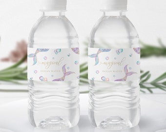 Op de afbeelding: Twee doorzichtige plastic waterflessen met witte doppen. Elke fles heeft een etiket met een zeemeerminstaartontwerp in tinten paars, blauw en teal, samen met het woord "magical" en "Ocean Water". De flessen staan op een wit bord.