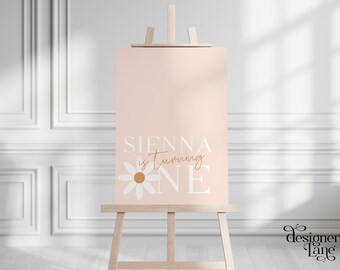 May include: A pink printable sign with the text "Sienna is turning one" in white with a white daisy with a brown center.
