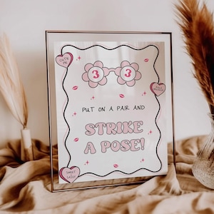 May include: A pink and white printable sign with the text "Put on a pair and strike a pose!"  The sign features a pair of pink glasses with the number 3 on each lens.  The sign is framed in a rose gold frame.