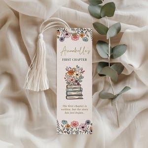 May include: A beige bookmark with a white tassel, featuring floral illustrations and text. The bookmark has a floral border and a stack of books with flowers on top. The text reads "Annabelle's FIRST CHAPTER" and "The first chapter is written, but the story has just begun."