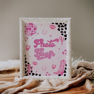 Könnte beinhalten: Ein weißes Fotobooth-Schild mit rosa Hintergrund und rosa und schwarzen Illustrationen. Das Schild trägt die Aufschrift "Photo Booth" in einer rosa Retro-Schrift.