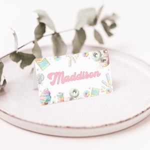 May include: White place card with a pink background and the name Maddison in pink script. The card is decorated with colourful donuts, ice cream cones, and other sweets.