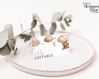 May include: A printable baby shower place card with a cute cartoon bear wearing a pilot's hat and goggles, flying in a hot air balloon. The card has a light brown and white color scheme with clouds and stars. The text "fully editable" is printed on the card.